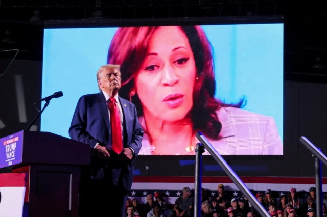 Donald Trump in blue suit stands on stage next to a podium. Behind him is a group of supporters. In the background is a maxi screen showing a close-up picture of Kamala Harris