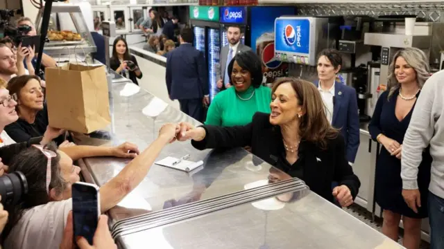 Kamala Harris fist bumping a customer at 4th Street Delicatessen in Philadelphia, Pennsylvania. She is in black pants suit and has an American pin on her left lapel. In the background are members of staff observing and the deli's kitchen is visible