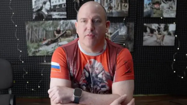 A man wearing a red t-shirt with a wolf on the front and a Russian flag on the sleeve, in front of a corkboard with pictures of scenes of military action