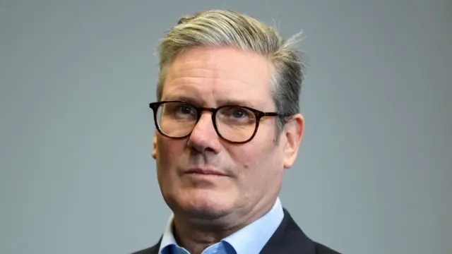 A headshot of Sir Keir Starmer shows him with glasses on, looking into the distance past the camera. He is wearing a black suit jacket with a light blue shirt