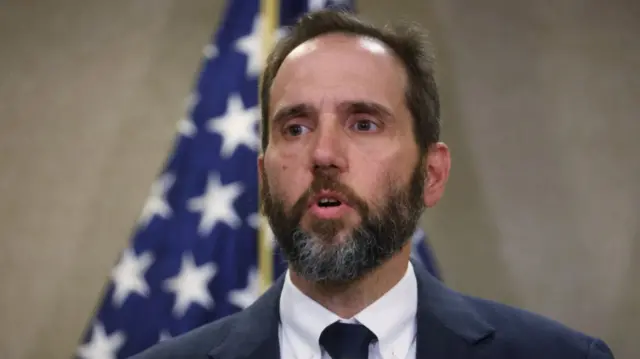 Special Counsel Jack Smith close up from top of the shoulders. He's in dark blue suit, white shirt and black tie. Beige wall and blurred US flag in the background