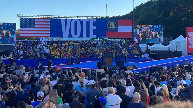 Spike Lee stands at a podium dressed in a burgundy tracksuit and thousands of people cheer in the crowd of a large football stadium