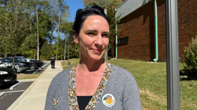 A woman stands in a grey sweater with a sticker on her left shoulder that shows she's voted. It has a peach on it