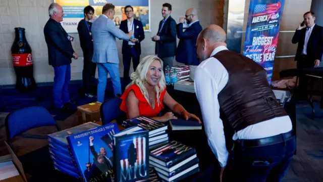 US Representative Marjorie Taylor Greene, in red top, signs books for supporters. She is speaking to a man in leather vest while sitting at a table her biography and a book about Trump on display. A group of men talks in the background