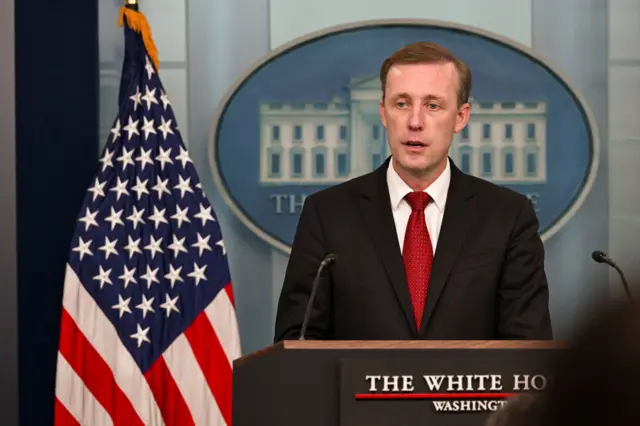 US National Security Advisor Jake Sullivan standing at a podium in black suit, white shirt and red patterned tie. White House Logo on blue background behind him