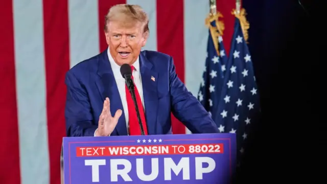 Donald Trump in dark blue suit, white shirt and red tie as he delivers speech. He has right hand raised as he leans his arm on the podium. A large US flag serves as background