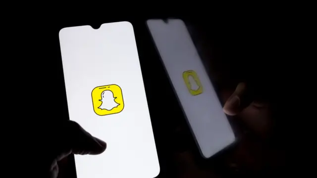 A hand holds a phone with the snapchat logo on the screen. The phone illuminates the hand, which is otherwise in darkness.