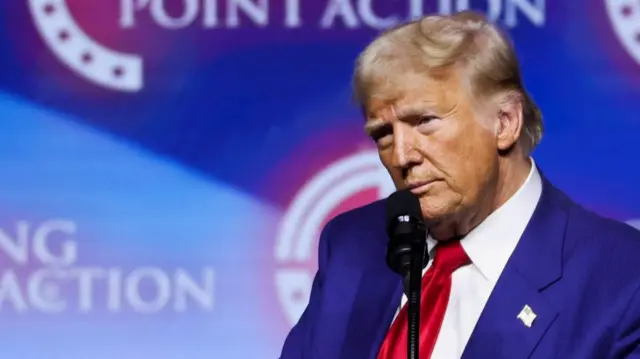 Donald Trump is seen in front of a blue background with the words “Turning Point Action.”