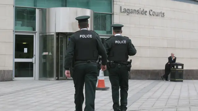 Two PSNI officers walk toward Laganside Courts, their backs to the camera. In the background, a man leans on a bin. There is an orange and white cone in front of the PSNI officers.