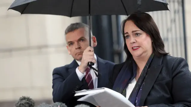 A woman and a man stand under an umbrella. The man is holding the umbrella, and the woman is holding a notebook. The man is wearing a dark suit with a maroon tie, while the woman is wearing a dark jacket. They are standing in front of several microphones.