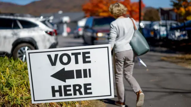 A 'vote here' sign, pointing left. Behind it, a blonde woman walks towards a car park.