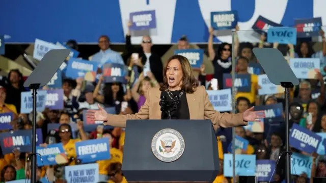 Kamala Harris stands on a podium, arms stretched out as she addresses the crowd. She is wearing a brown jacket and black top