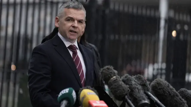 A man in a dark suit and maroon tie stands in front of several microphones. He has grey hair.