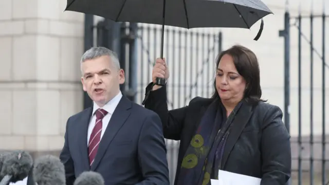 A man in a dark suit with a maroon tie and a woman in a dark jacket stand next to each other. The man has grey hair, while the woman has dark hair and is holding a black umbrella. The man in standing in front of several grey microphones.