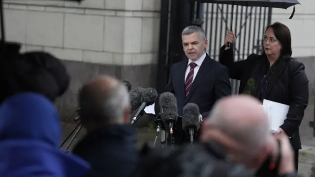 A man with grey hair and a woman with dark hair stand in front of several microphones. The man is wearing a dark suit with a maroon tie. The woman is wearing a dark jacket and is holding a black umbrella. In the foreground, the backs of numerous people's heads can be seen, out of focus.