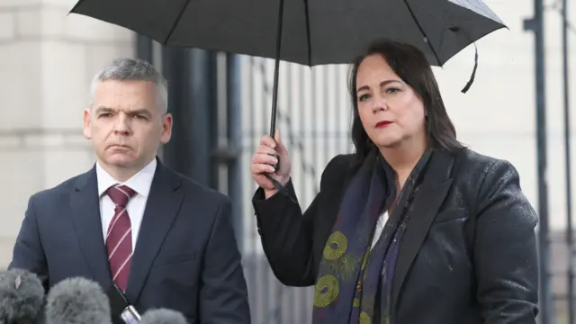 A man in a dark suit and a woman in a dark jacket stand under a black umbrella in front of several grey microphones.