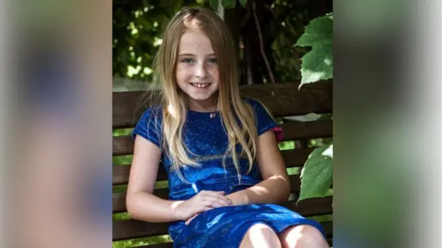 Cimarron sitting on a park bench, hands folded and wearing a blue dress