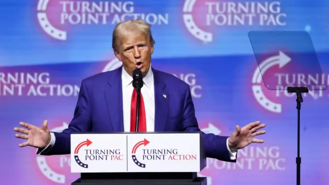 Donald Trump stands on a podium with his arms out as he addresses the crowd at a rally. He is wearing a blue suit, white shirt and red tie