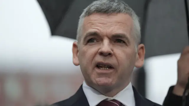 A close-up of a man with grey hair, wearing a dark suit. Behind him, a hand holds a black umbrella.