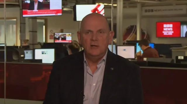 A man speaking to a camera in front of a newsroom