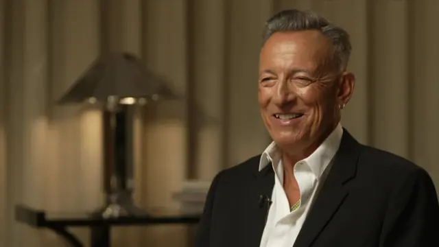 Bruce Springsteen can be seen in a black suit with a white shirt on, smiling at the interview who is out of camera shot