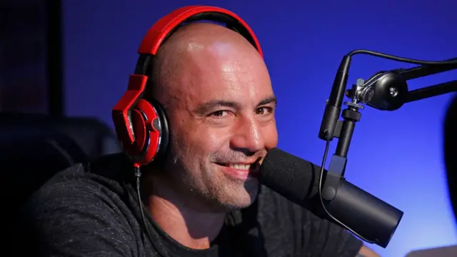 Joe Rogan, wearing orange headphones, smiles into a microphone during a taping of Joe Rogan Questions Everything