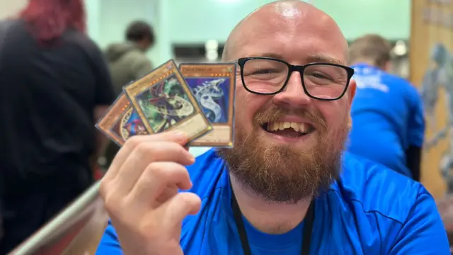 James Tiffin, a bald man with ginger beard and black-rimmed glasses, smiles as he holds up three YuGi-Oh cards