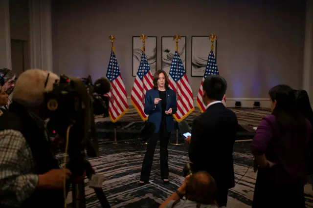 Harris stands in front of four American flags with reporters in the foreground