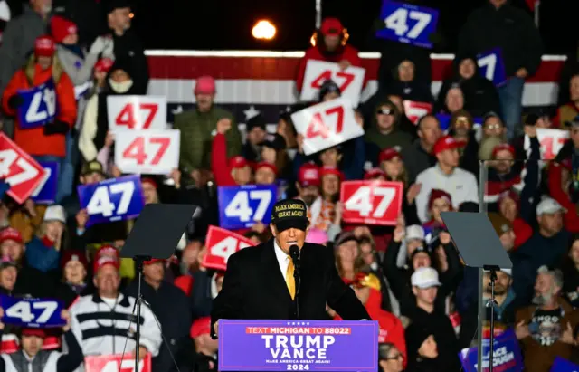 Trump wears a black hat while supporters wave signs that read 47