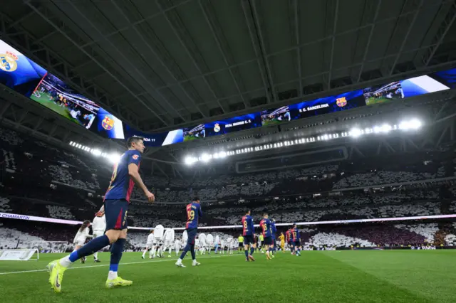 The players make their way out onto the pitch as the fans hold up a tifo in the stands