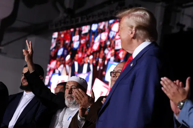 A Muslim community leaders holds two fingers up in a "peace sign" next to Donald Trump on stage in Michigan