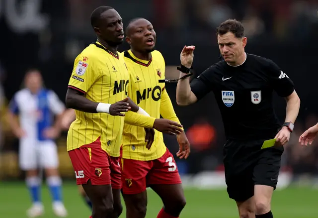 Referee Darren England (right) shows Watford's Edo Kayembe (centre) a yellow card