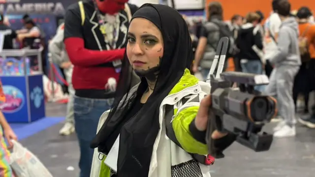 Cosplayer Dani known online as Hijabi Hoo dressed as Crypto from Apex Legends with a white and neon jacket, holding a fake gun