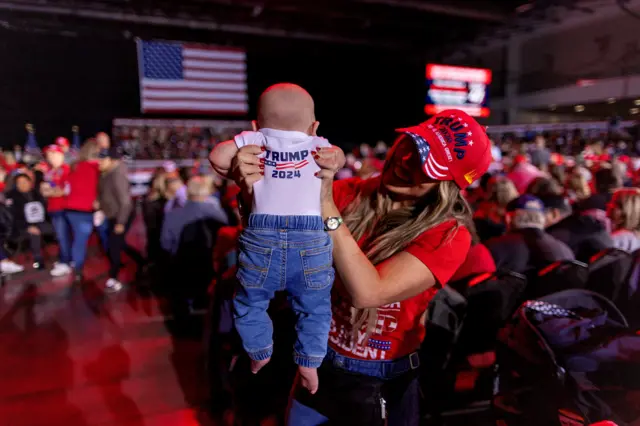 A woman holds up her baby who is dressed in Trump merch at a rally in Michigan