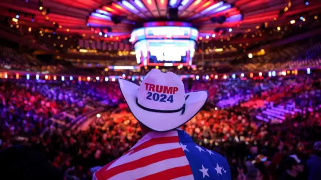 Someone stands looking away from the camera wearing a cowboy hat with slogan 'Trump 2024'. The rally crowd can be seen in background