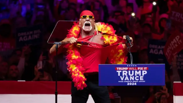 Hulk Hogan stands at a Trump podium and tears off his red tank top. He is wearing a red and yellow feathe boa and yellow sunglasses with a red hat.