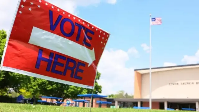 Sign saying 'Vote here' on a lawn in front of a building