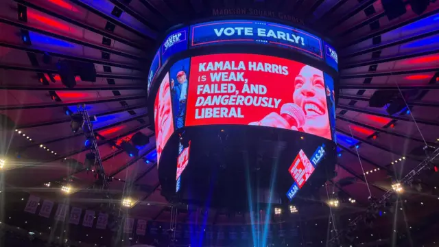 A jumbotron shows an attack ad against Kamala Harris which calls her weak, failed and dangerously liberal