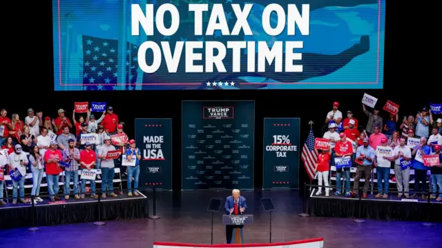 Donald Trump speaks on stage with a big sign above his head, which reads: "No tax on overtime"