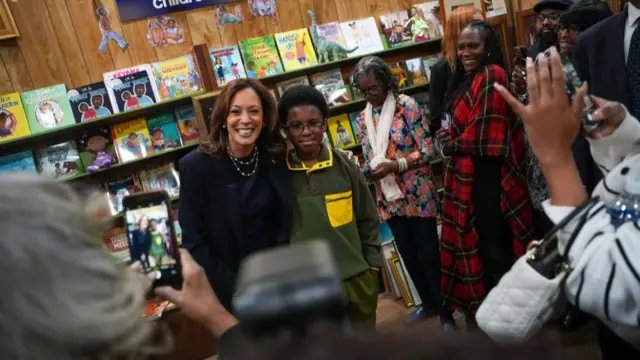 Harris smiles with her around a young black boy in a green sweatshirt as photographers take pictures