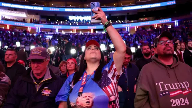 A woman in a trump shirt takes a picture inside Madison Square Garden ahead of the Donald Trump rally. She's surrounded by men wearing Trump hats and Trump shirts