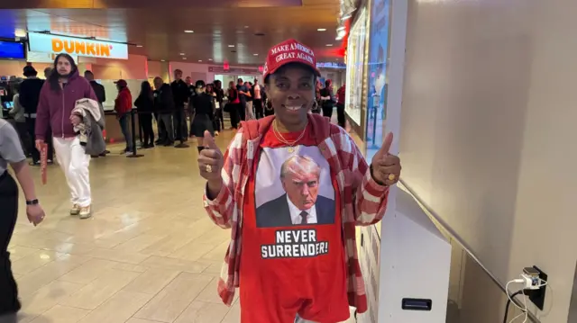 A woman stands in a red T-shirt with Donald Trump's mugshot picture, and the words, "Never surrender" written below