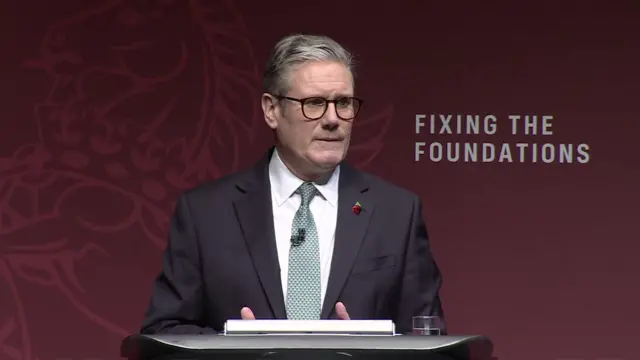 Keir Starmer delivers speech in Birmingham. He's wearing black suit with white blouse and light turquoise patterned tie. He has poppy pin on his left lapel and is wearing glasses. Background is a burgundy red panel with Fixing the Foundations written on it