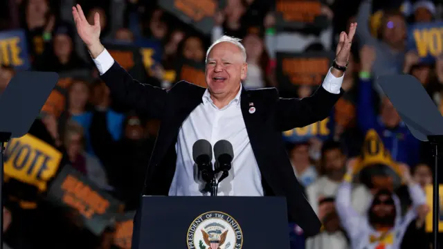 Tim Walz grins as he raises his arms on stage in Michigan