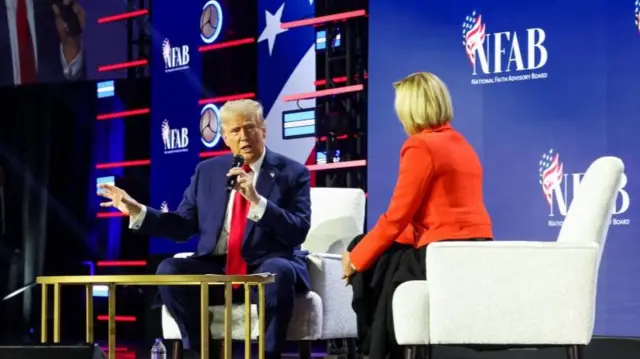 Trump sitting at a chair speaking into a microphone facing the camera. A woman sits next to him facing away from the camera looking towards trump