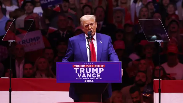 Trump on stage and speaking at the podium at Madison Square Garden, with part of the crowd shown behind him