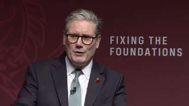 Mid-shot picture of Keir Starmer delivering speech. He's in black suit, white blouse and patterned turquoise tie. Poppy pin on his left lapel and he's wearing glasses. Burgundy background