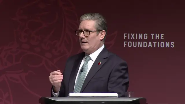 Keir Starmer delivers speech in black suit, white shirt and patterned tie. His right hand is raised and partially cupped as he answers questions