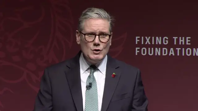 Keir Starmer in black suit, white shirt and patterned turquoise tie, poppy pin on left lapel. He's wearing black glases and open is partly open as he speaks