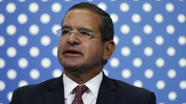 Pedro Pieruluisi, wearing glasses, black blazer, white shirt and a patterned tie, against a blue and white polka dot background